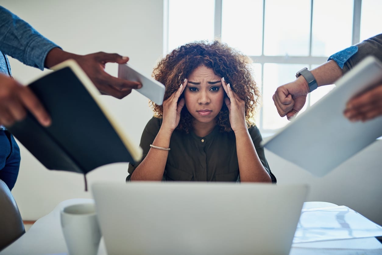 Photo d'une femme se tenant la tête, entourée de dossiers tendues vers elle au travail 